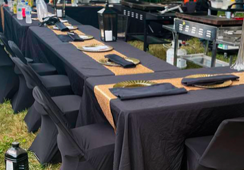 Outdoor Dining Setup With Black Clothed Tables, Place Settings, And A Food Station In The Background With A Staff Member Wearing A Mask.